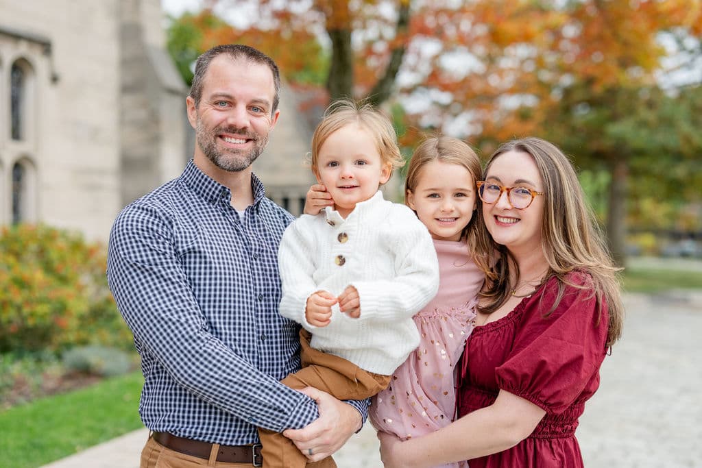 Family smiling together during an outdoor fall mini photo session in Pittsburgh, with colorful autumn trees in the background